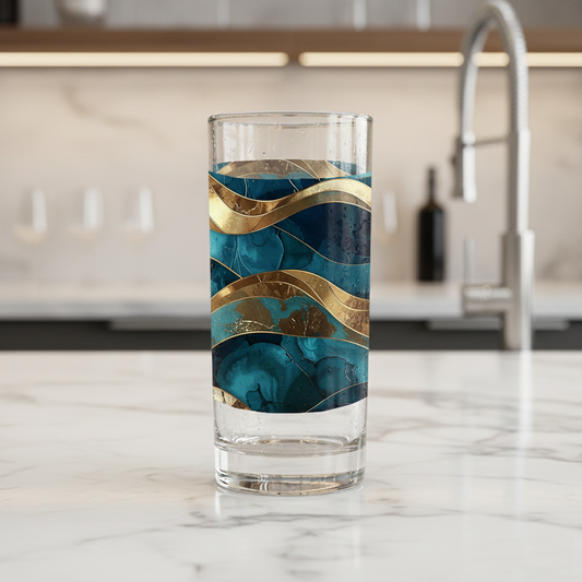 Opulent Teal highball glass with teal and gold wavy design on a marble countertop in a kitchen with blurred background.