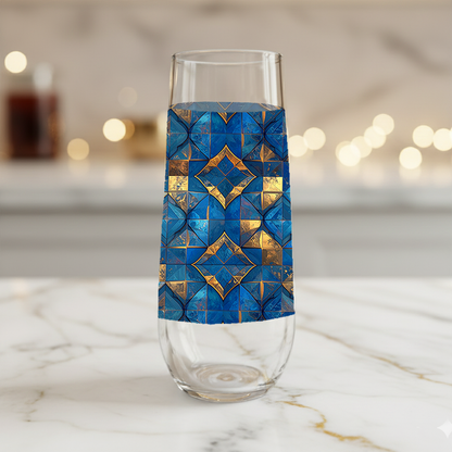 Moroccan Treasure stemless champagne glass with blue and gold geometric design on a marble surface and blurred lights in background.
